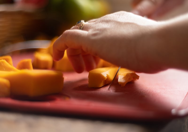 Hands chopping pumpkin into one-inch pieces on a red cutting board.