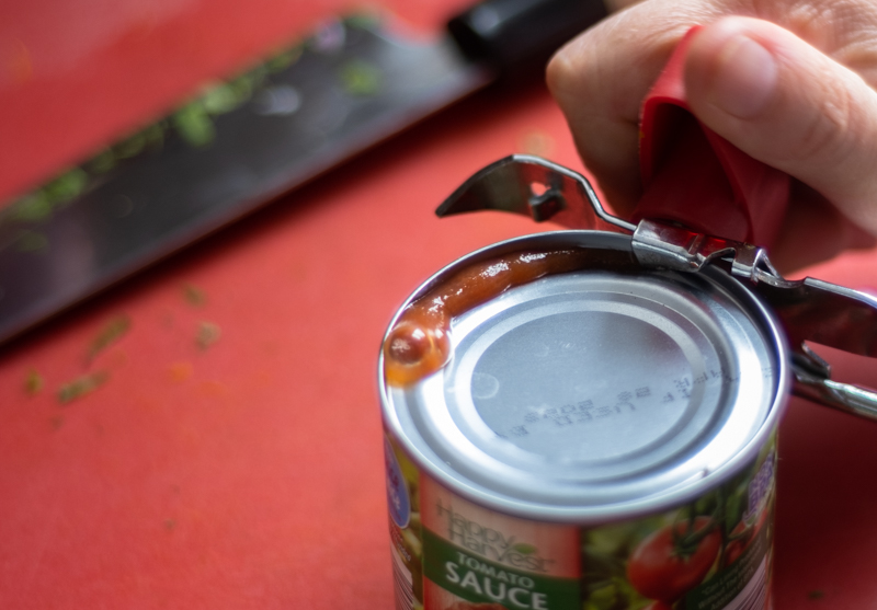 Hands using a can opener to open a can of tomato sauce.