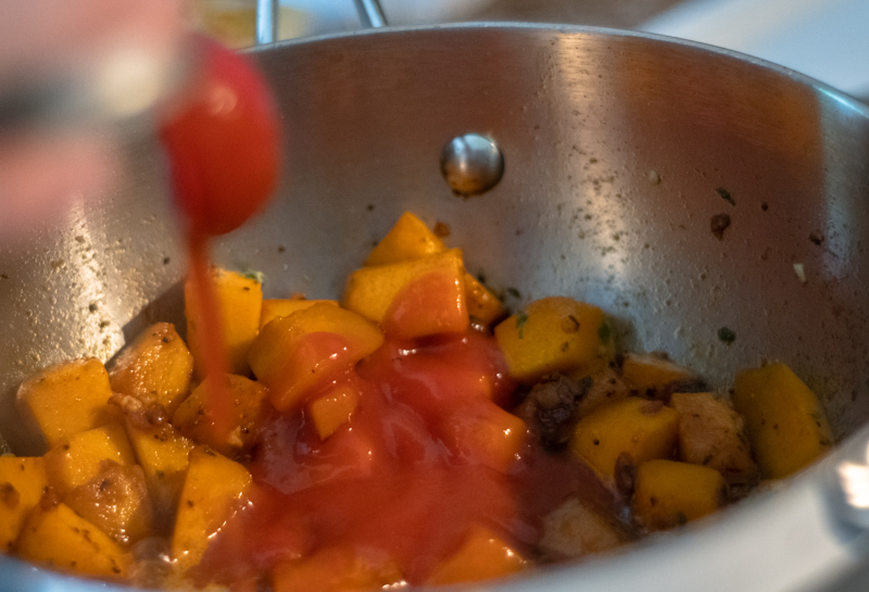 Tomato sauce being added to pumpkin, purple onion, and spices in a pot.